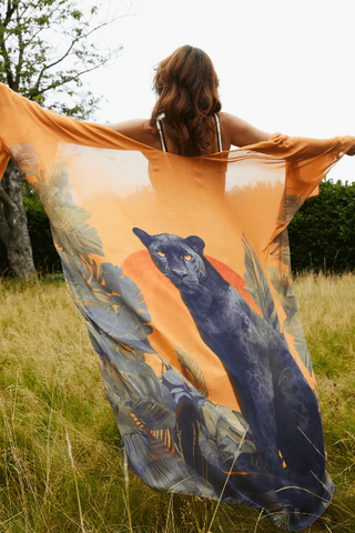 Woman holding a large orange scarf with a panther design in a grassy field.