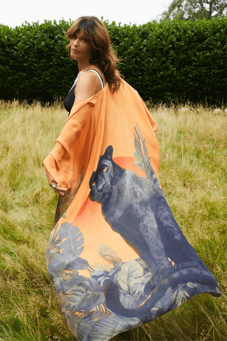 Woman holding an orange scarf with a panther design in a grassy field.