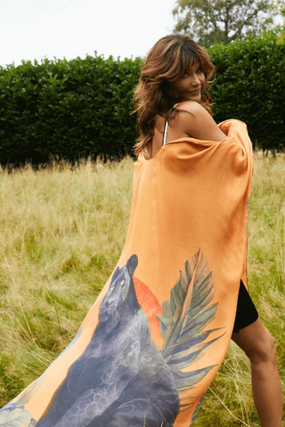 Woman standing in a field with a large orange towel draped over her shoulders featuring a bird and leaf design.